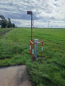 rotweißes Gestell, das auf einer Wiese mit Windrädern im Hintergrund eine Grundwassermessstelle eingrenzt und mittels einer Fahne markiert
