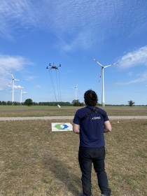 Ein Mitarbeiter der Firma ASDRO steht im Vordergrund und steuert eine Drohne über einer landwirtschaftlichen Fläche. Im Hintergrund sind Windräder zu sehen.