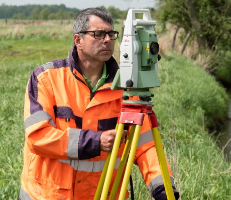Vermessungsarbeiten an einem Bach auf einem Feld. Im Vordergrund  schaut ein Mann in Arbeitskleidung durch ein Vermessungsgerät.